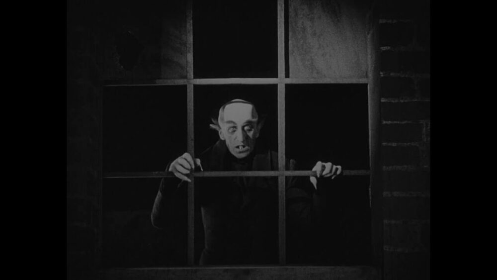 暗い窓枠の向こうから細い指で柵をつかみ、表情を強張らせた人物がこちらを見つめている様子。